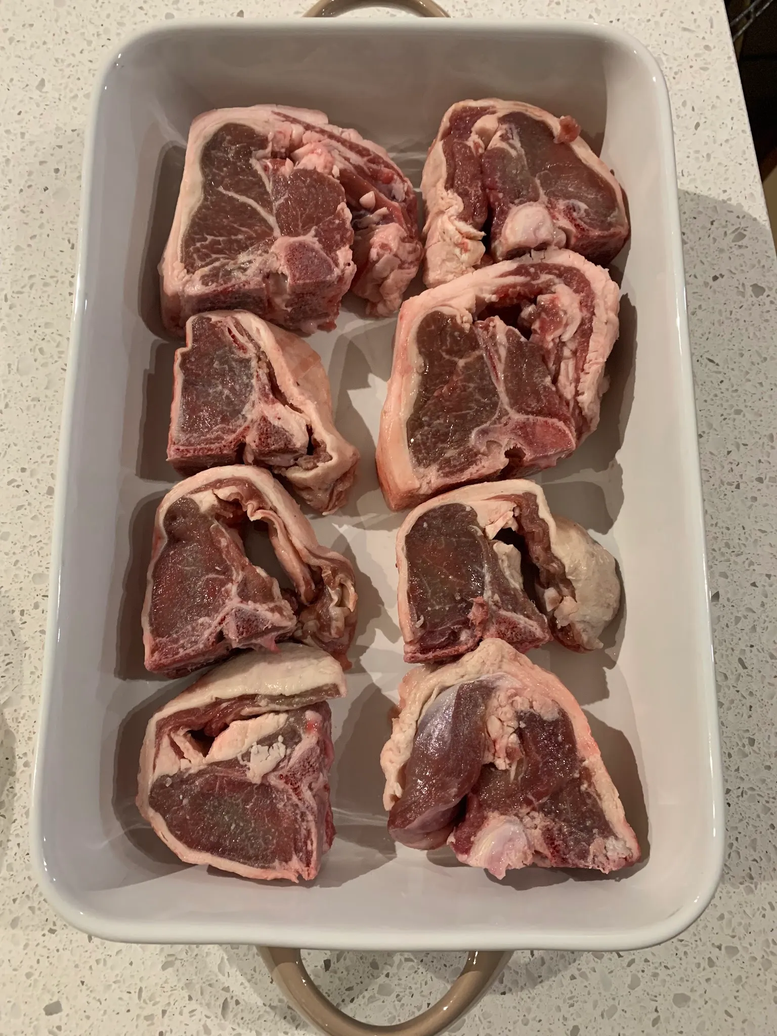 Eight raw lamb chops arranged neatly in a white ceramic baking dish, shot from above on a white benchtop
