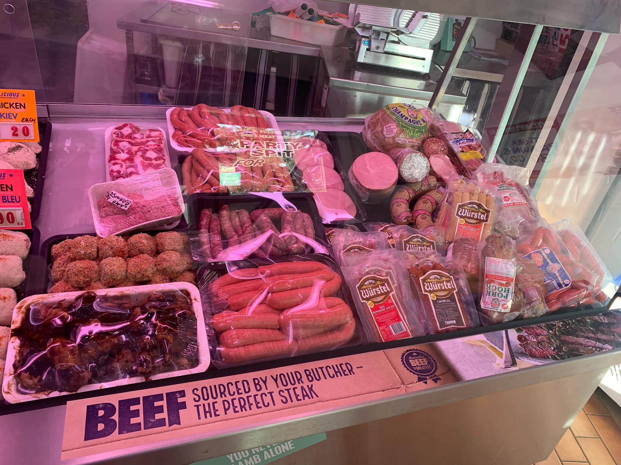 Wide butcher display case section showing sausages, meatballs, marinated skewers, and packaged smallgoods under pink shop lighting
